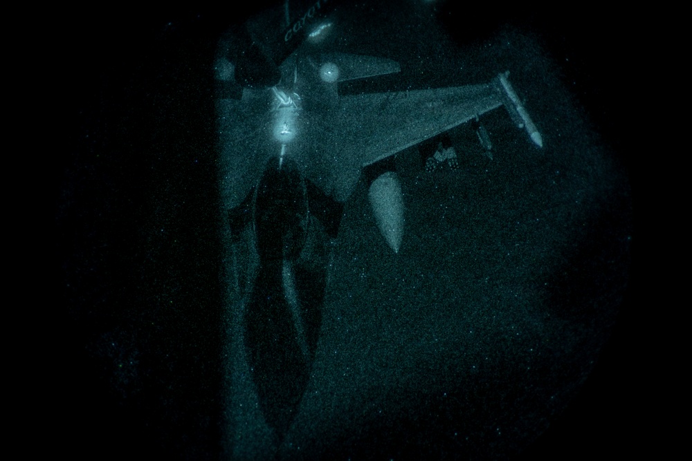 A night-vision or low-light image capturing what appears to be an aerial refueling operation between two military aircraft. The image shows a fighter jet connected to a tanker aircraft via a refueling boom or drogue system. The grainy, teal-tinted appearance is consistent with night-vision goggle (NVG) photography.