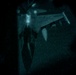 A night-vision or low-light image capturing what appears to be an aerial refueling operation between two military aircraft. The image shows a fighter jet connected to a tanker aircraft via a refueling boom or drogue system. The grainy, teal-tinted appearance is consistent with night-vision goggle (NVG) photography.