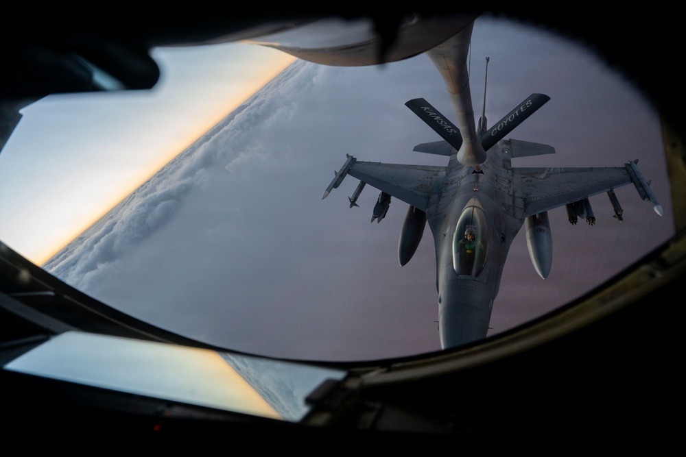 An aerial refueling operation is captured from the boom operator's window of a tanker aircraft, showing an F-16 Fighting Falcon receiving fuel via a refueling boom. The image is taken from below/behind the tanker looking down at the receiver aircraft against a twilight sky with cloud cover below. Text on the refueling boom reads 'KANSAS COYOTES', indicating a Kansas Air National Guard unit.