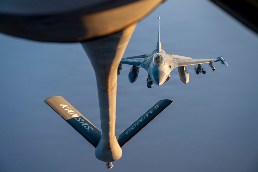 An aerial refueling operation is captured from the perspective of a tanker aircraft, showing a fighter jet approaching the refueling boom. The tanker's boom is prominently visible in the foreground with 'KANSAS' and 'COYOTES' markings on its stabilizer fins. The fighter aircraft, identifiable as an F-16 Fighting Falcon, is positioned directly behind the boom in a pre-contact or contact refueling position.