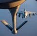 An aerial refueling operation is captured from the perspective of a tanker aircraft, showing a fighter jet approaching the refueling boom. The tanker's boom is prominently visible in the foreground with 'KANSAS' and 'COYOTES' markings on its stabilizer fins. The fighter aircraft, identifiable as an F-16 Fighting Falcon, is positioned directly behind the boom in a pre-contact or contact refueling position.