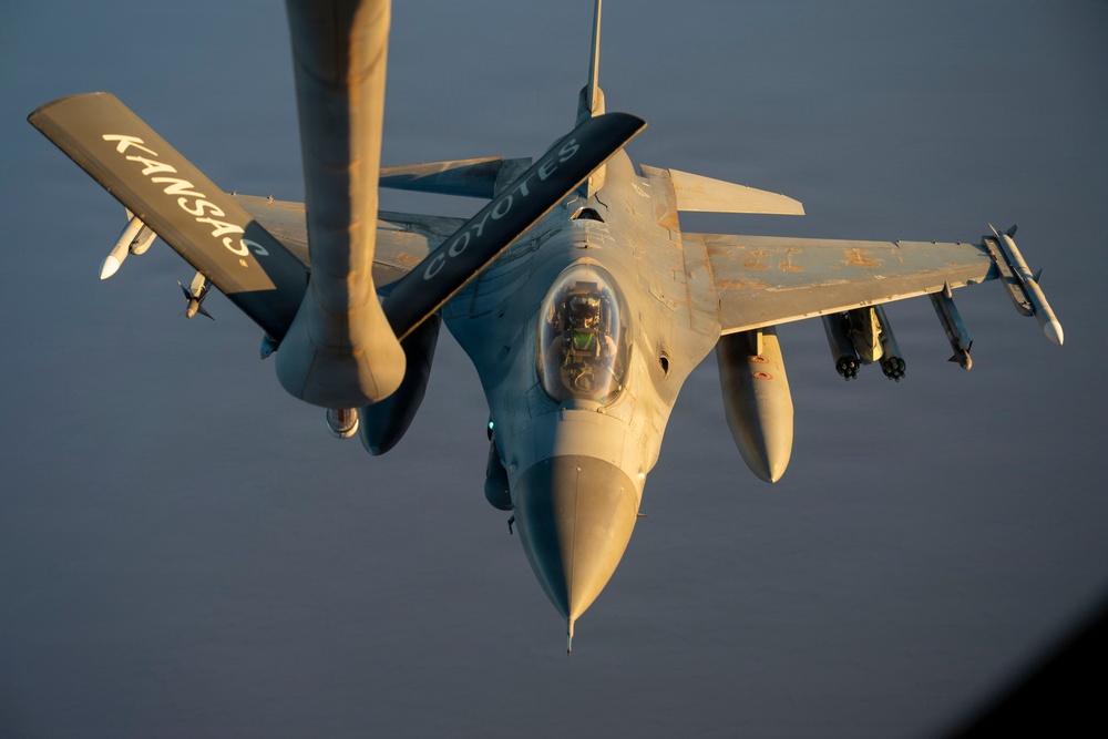 An aerial photograph captures an F-16 Fighting Falcon jet fighter during an in-flight refueling operation. The aircraft bears the markings 'KANSAS' on the wing and 'COYOTES' on the fuselage, indicating it belongs to a Kansas Air National Guard unit. The image is taken from the perspective of the tanker aircraft, with the refueling boom visible in the foreground.