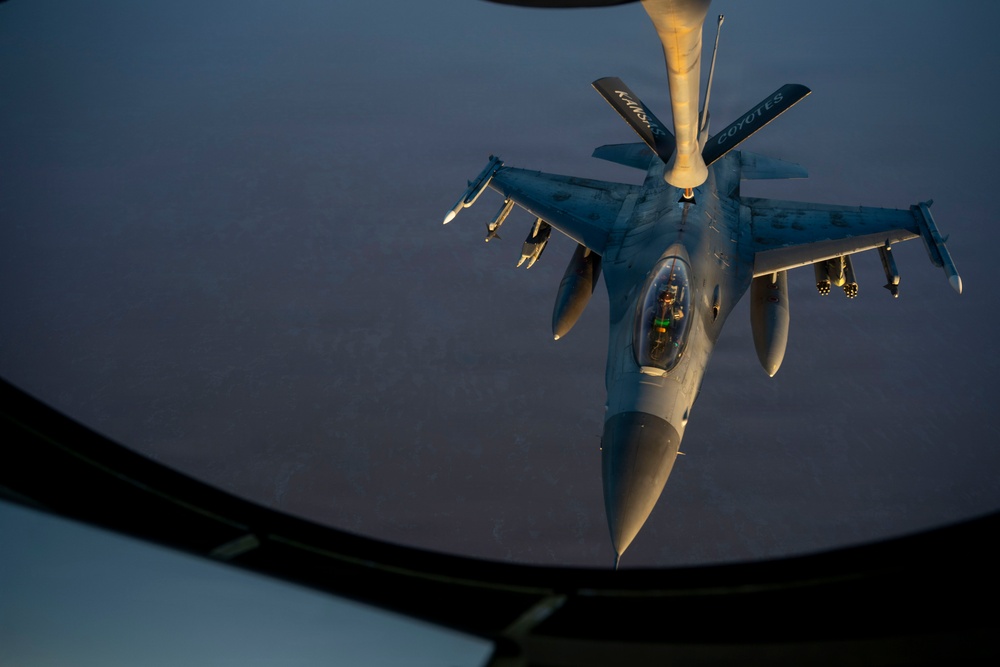 An aerial refueling operation is captured from the boom operator's window of a tanker aircraft, showing an F-16 Fighting Falcon receiving fuel via a refueling boom. The image is taken at dusk or dawn, with warm golden light illuminating the fighter jet against a dark blue sky. The word 'COYOTES' is visible on the refueling boom, suggesting a unit designation.