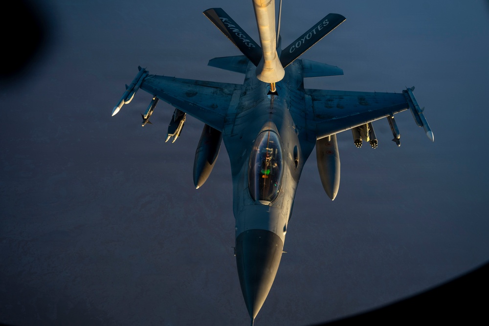 An F-16 Fighting Falcon jet fighter is receiving aerial refueling from a tanker aircraft, photographed from above during low-light conditions at dusk or dawn. The refueling boom from the tanker, marked with 'KANSAS COYOTES,' is connected to the fighter's receptacle. The aircraft is fully armed with multiple munitions visible on its wing pylons.
