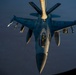 An F-16 Fighting Falcon jet fighter is receiving aerial refueling from a tanker aircraft, photographed from above during low-light conditions at dusk or dawn. The refueling boom from the tanker, marked with 'KANSAS COYOTES,' is connected to the fighter's receptacle. The aircraft is fully armed with multiple munitions visible on its wing pylons.