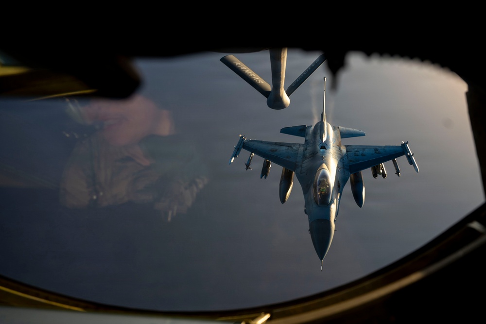 An aerial refueling operation is captured from inside a tanker aircraft, showing an F-16 Fighting Falcon approaching the refueling boom. The image is taken through the boom operator's window of what appears to be a KC-135 Stratotanker, with the refueling boom extended toward the fighter jet. The scene is photographed during low-light conditions, likely at dusk or dawn, casting a warm golden hue across the aircraft.