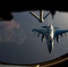 An aerial refueling operation is captured from inside a tanker aircraft, showing an F-16 Fighting Falcon approaching the refueling boom. The image is taken through the boom operator's window of what appears to be a KC-135 Stratotanker, with the refueling boom extended toward the fighter jet. The scene is photographed during low-light conditions, likely at dusk or dawn, casting a warm golden hue across the aircraft.