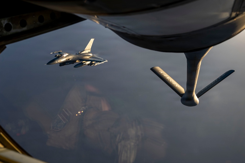 An aerial refueling operation is captured from the perspective of a tanker aircraft, showing its refueling boom extended in the foreground. A fighter jet, identifiable as an F-16 Fighting Falcon, is positioned nearby in formation, likely awaiting or completing fuel transfer. The image is taken at low sun angle, casting warm golden light across the scene with terrain visible far below.