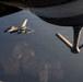 An aerial refueling operation is captured from the perspective of a tanker aircraft, showing its refueling boom extended in the foreground. A fighter jet, identifiable as an F-16 Fighting Falcon, is positioned nearby in formation, likely awaiting or completing fuel transfer. The image is taken at low sun angle, casting warm golden light across the scene with terrain visible far below.