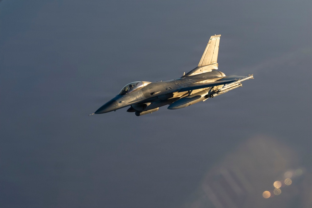 A single-engine fighter jet, consistent with an F-16 Fighting Falcon design, is captured in flight against an overcast gray sky. The aircraft is illuminated by warm golden light, suggesting either sunrise or sunset conditions. Several munitions or external stores are visible mounted under the wings.