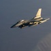 A single-engine fighter jet, consistent with an F-16 Fighting Falcon design, is captured in flight against an overcast gray sky. The aircraft is illuminated by warm golden light, suggesting either sunrise or sunset conditions. Several munitions or external stores are visible mounted under the wings.