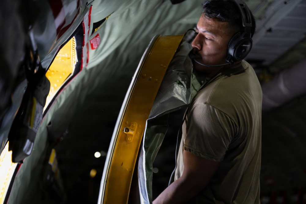 A military or aviation personnel member wearing olive/tan military-style clothing and Bose headphones is inspecting or operating a large aircraft door or hatch mechanism. The individual appears to be inside a large military transport aircraft, examining a door frame with a distinctive yellow/gold-colored seal or gasket. The lighting is dramatic, with warm yellow light emanating from the door opening contrasting with the darker interior.