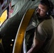 A military or aviation personnel member wearing olive/tan military-style clothing and Bose headphones is inspecting or operating a large aircraft door or hatch mechanism. The individual appears to be inside a large military transport aircraft, examining a door frame with a distinctive yellow/gold-colored seal or gasket. The lighting is dramatic, with warm yellow light emanating from the door opening contrasting with the darker interior.