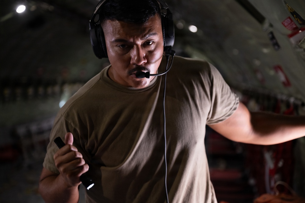 A military or aviation crew member is photographed inside what appears to be a military cargo aircraft. He is wearing a communications headset with a boom microphone and an olive drab t-shirt, and appears to be actively communicating or directing operations. He holds a small flashlight or tool in one hand while gesturing with the other.