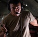 A military or aviation crew member is photographed inside what appears to be a military cargo aircraft. He is wearing a communications headset with a boom microphone and an olive drab t-shirt, and appears to be actively communicating or directing operations. He holds a small flashlight or tool in one hand while gesturing with the other.