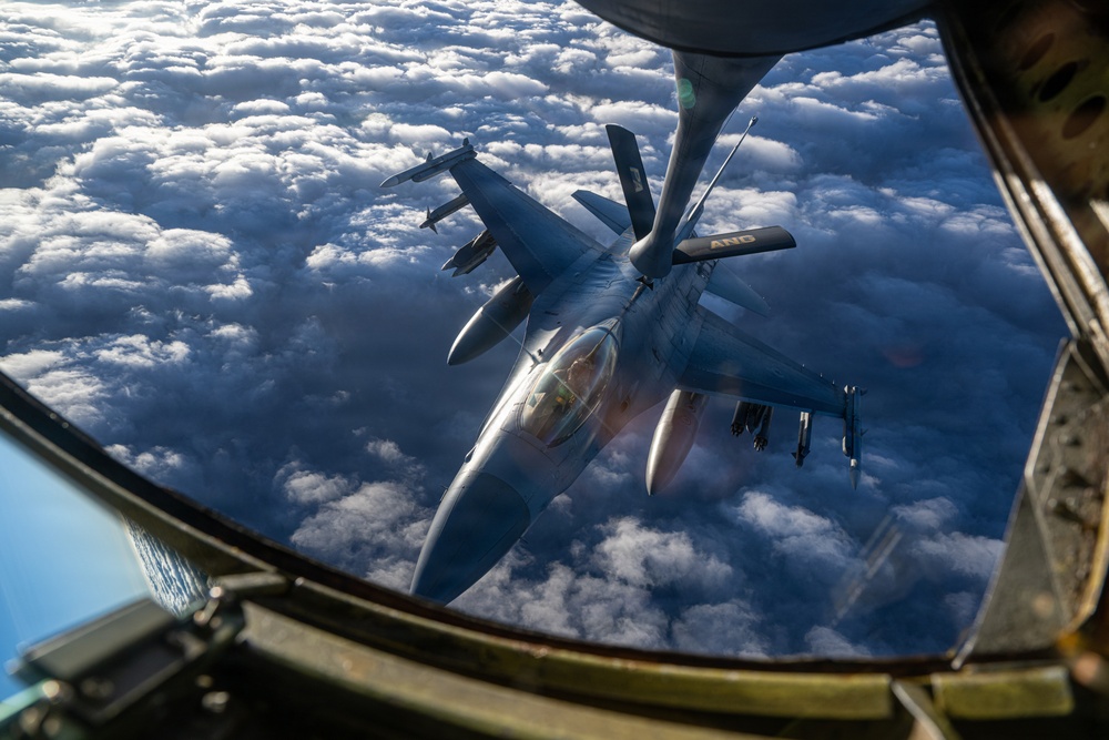 An aerial refueling operation is captured from the boom operator's station of a tanker aircraft, showing an F-16 Fighting Falcon receiving fuel via a flying boom. The fighter jet is photographed from above and behind, with a dense cloud layer visible far below. Text reading 'ANG' (Air National Guard) is visible on the aircraft's tail fin.