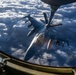An aerial refueling operation is captured from the boom operator's station of a tanker aircraft, showing an F-16 Fighting Falcon receiving fuel via a flying boom. The fighter jet is photographed from above and behind, with a dense cloud layer visible far below. Text reading 'ANG' (Air National Guard) is visible on the aircraft's tail fin.