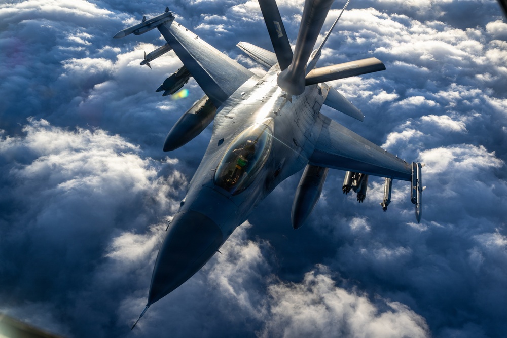 An aerial photograph captures an F-16 Fighting Falcon jet fighter receiving in-flight refueling from a tanker aircraft via a boom refueling system. The image is taken from above and slightly forward, likely from the tanker aircraft itself, showing the refueling boom connected to the fighter's receptacle. A second F-16 is visible in the background, and the aircraft are flying above a dense cloud layer.