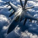 An aerial photograph captures an F-16 Fighting Falcon jet fighter receiving in-flight refueling from a tanker aircraft via a boom refueling system. The image is taken from above and slightly forward, likely from the tanker aircraft itself, showing the refueling boom connected to the fighter's receptacle. A second F-16 is visible in the background, and the aircraft are flying above a dense cloud layer.