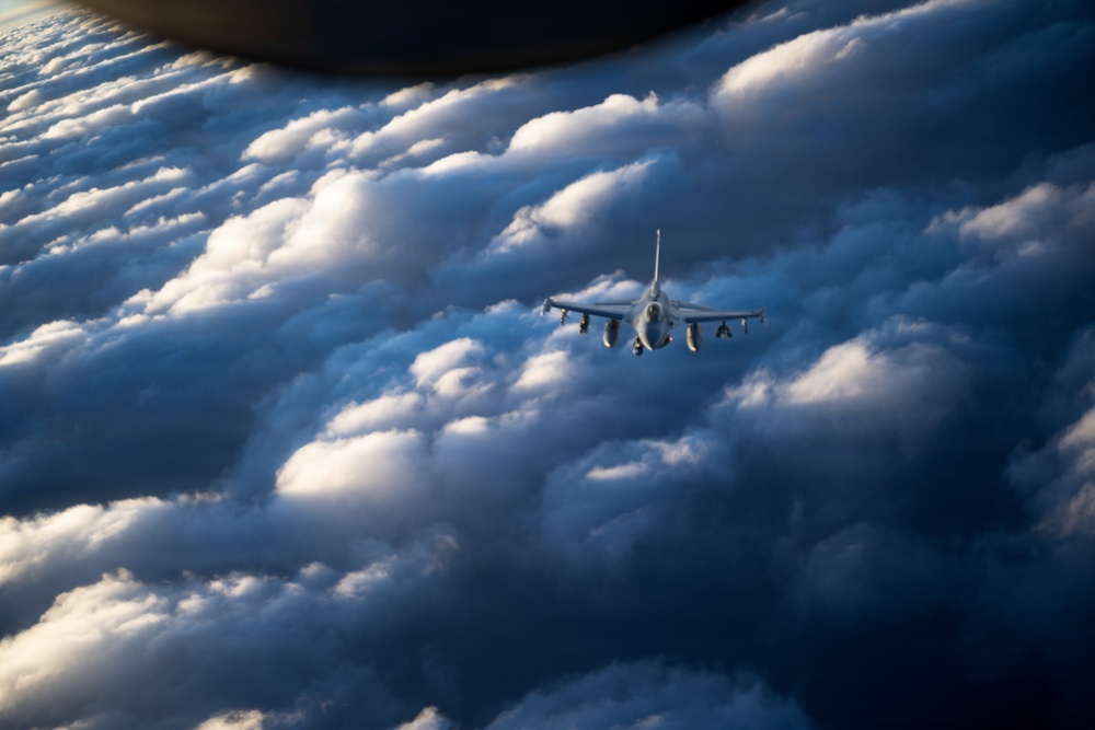 A military fighter jet is photographed in flight above a dramatic cloudscape, likely taken from another aircraft. The jet appears to be flying at high altitude among large cumulus clouds illuminated by directional sunlight. The perspective suggests an air-to-air photography scenario, possibly during a formation flight or escort mission.