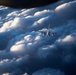 A military fighter jet is photographed in flight above a dramatic cloudscape, likely taken from another aircraft. The jet appears to be flying at high altitude among large cumulus clouds illuminated by directional sunlight. The perspective suggests an air-to-air photography scenario, possibly during a formation flight or escort mission.