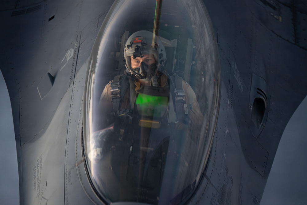 A military fighter pilot is seated inside the cockpit of what appears to be an F-16 Fighting Falcon, photographed through the bubble canopy. The pilot is wearing full flight gear including a helmet with night vision goggles mounted, an oxygen mask, and a flight suit with harness straps. A green HUD (Heads-Up Display) reflection is visible on the canopy glass, suggesting the aircraft's avionics are active.