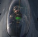 A military fighter pilot is seated inside the cockpit of what appears to be an F-16 Fighting Falcon, photographed through the bubble canopy. The pilot is wearing full flight gear including a helmet with night vision goggles mounted, an oxygen mask, and a flight suit with harness straps. A green HUD (Heads-Up Display) reflection is visible on the canopy glass, suggesting the aircraft's avionics are active.