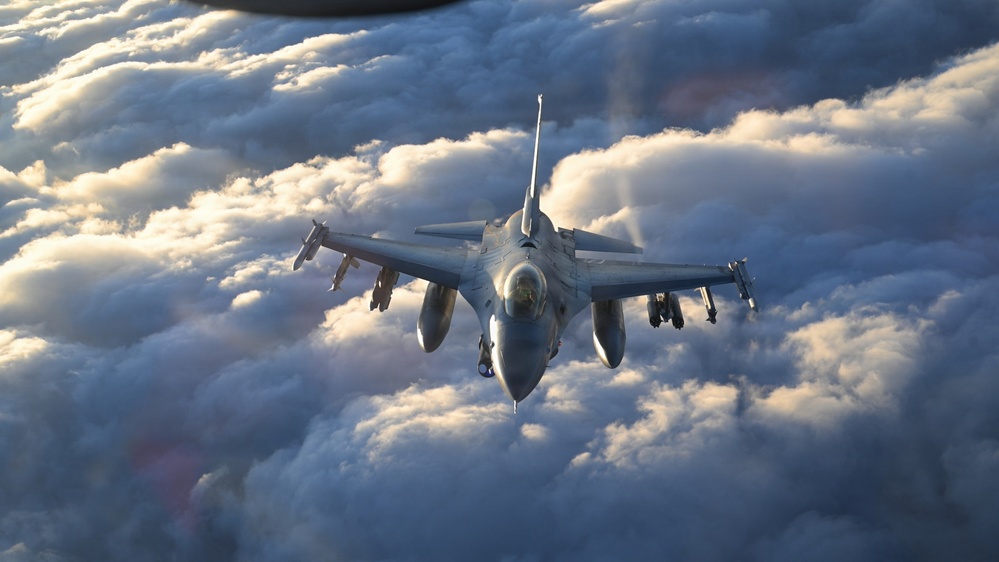 A single-engine fighter jet, identifiable as an F-16 Fighting Falcon, is photographed head-on while flying above a dense cloud layer. The aircraft is carrying multiple wing-mounted stores including missiles and external fuel tanks. The image is taken from another aircraft, likely during a formation flight or aerial refueling mission, with warm golden light suggesting dawn or dusk conditions.