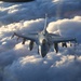 A single-engine fighter jet, identifiable as an F-16 Fighting Falcon, is photographed head-on while flying above a dense cloud layer. The aircraft is carrying multiple wing-mounted stores including missiles and external fuel tanks. The image is taken from another aircraft, likely during a formation flight or aerial refueling mission, with warm golden light suggesting dawn or dusk conditions.