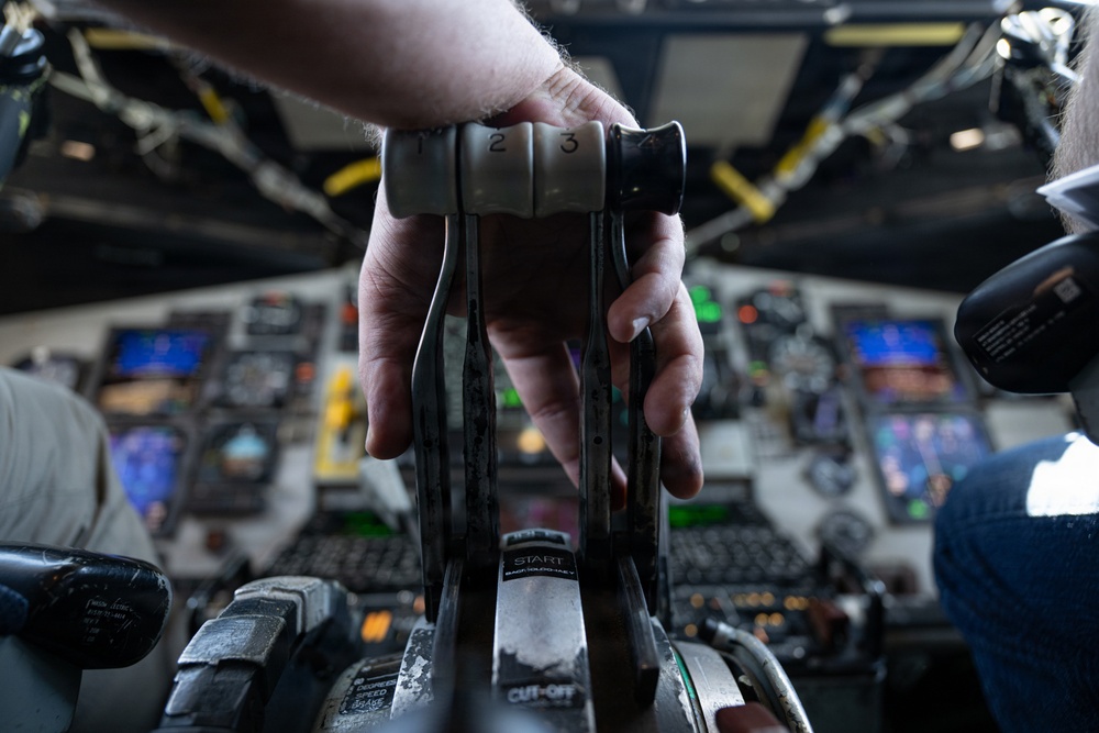 A close-up photograph of a pilot's hand gripping multiple throttle levers in a large aircraft cockpit, with levers numbered 1, 2, and 3 visible. Labels reading 'START' and 'CUT-OFF' are visible on the throttle quadrant. The background shows a complex instrument panel with multiple digital displays and analog gauges, consistent with a large commercial or military transport aircraft.