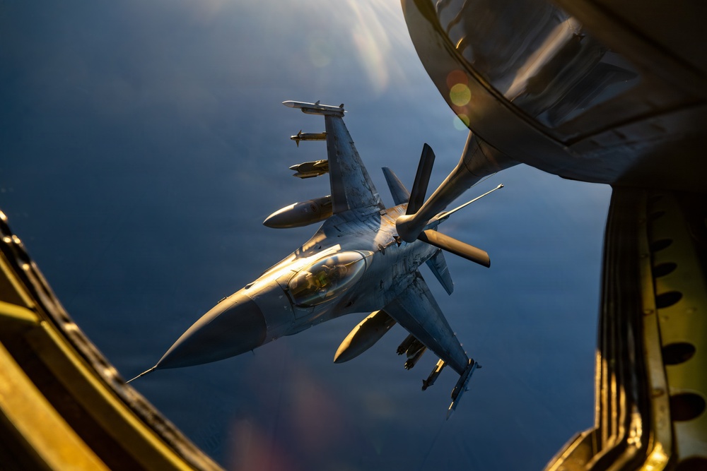 An F-16 Fighting Falcon jet fighter is captured mid-air during an aerial refueling operation, photographed from the boom operator's station of a tanker aircraft. The fighter is armed with multiple munitions on its wing pylons and is positioned close to the refueling boom extending from the tanker. The image is taken during golden hour lighting conditions, casting warm tones against a deep blue sky backdrop.