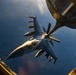An F-16 Fighting Falcon jet fighter is captured mid-air during an aerial refueling operation, photographed from the boom operator's station of a tanker aircraft. The fighter is armed with multiple munitions on its wing pylons and is positioned close to the refueling boom extending from the tanker. The image is taken during golden hour lighting conditions, casting warm tones against a deep blue sky backdrop.