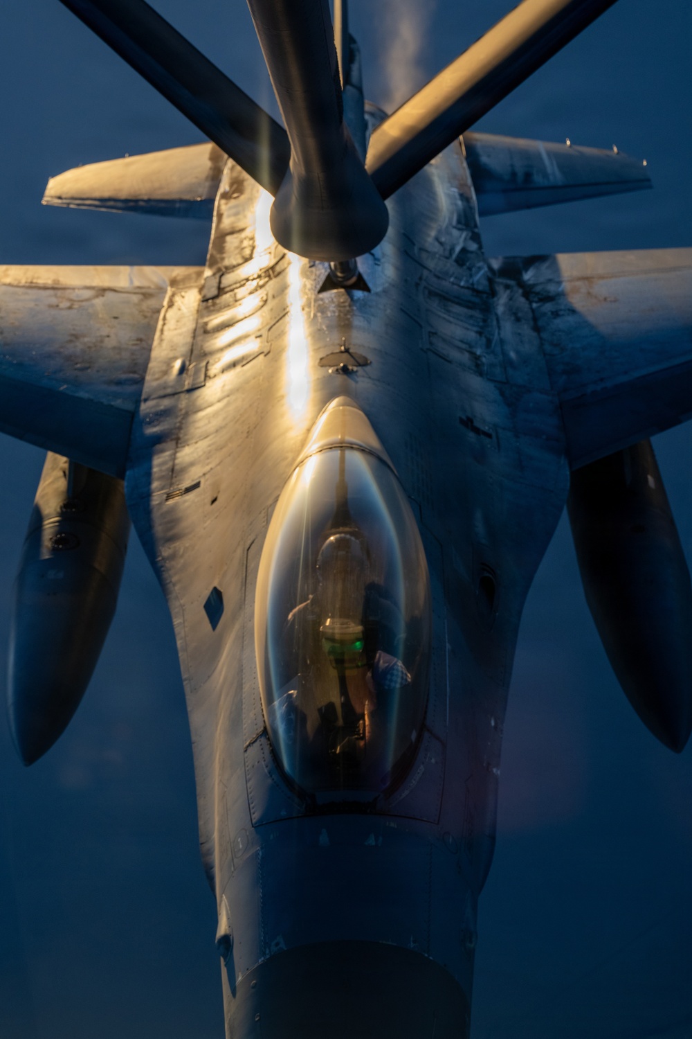 An aerial refueling operation is captured from the perspective of the tanker aircraft looking down at the receiver aircraft. The image shows a close-up top-down view of what appears to be a B-1B Lancer strategic bomber making contact with a refueling boom. The scene is illuminated by warm golden light, likely from a low-angle sun at dawn or dusk, contrasting with the deep blue sky.