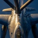 An aerial refueling operation is captured from the perspective of the tanker aircraft looking down at the receiver aircraft. The image shows a close-up top-down view of what appears to be a B-1B Lancer strategic bomber making contact with a refueling boom. The scene is illuminated by warm golden light, likely from a low-angle sun at dawn or dusk, contrasting with the deep blue sky.