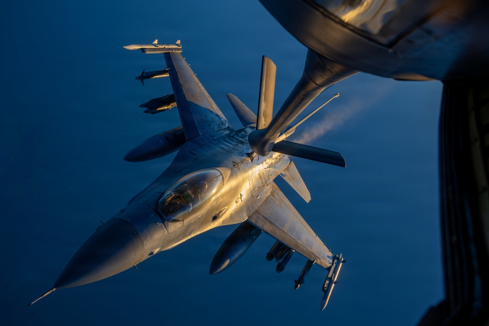 An F-16 Fighting Falcon jet fighter is captured in flight during an aerial refueling operation, photographed from the tanker aircraft above. The fighter is fully armed with multiple air-to-air and air-to-ground munitions visible on its wing pylons. The image is taken during golden hour lighting conditions at high altitude, with a deep blue sky visible in the background.