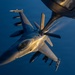 An F-16 Fighting Falcon jet fighter is captured in flight during an aerial refueling operation, photographed from the tanker aircraft above. The fighter is fully armed with multiple air-to-air and air-to-ground munitions visible on its wing pylons. The image is taken during golden hour lighting conditions at high altitude, with a deep blue sky visible in the background.