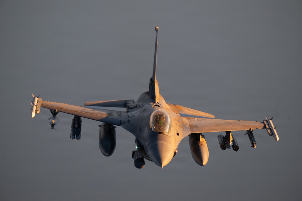 A front-facing view of an F-16 Fighting Falcon multirole fighter jet in flight, illuminated by warm golden-hour sunlight against an overcast gray sky. The aircraft is captured in a slightly nose-down attitude with landing gear deployed, suggesting it may be on approach or recently airborne. Multiple air-to-air missiles and external stores are visible on the wing hardpoints.
