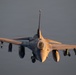 A front-facing view of an F-16 Fighting Falcon multirole fighter jet in flight, illuminated by warm golden-hour sunlight against an overcast gray sky. The aircraft is captured in a slightly nose-down attitude with landing gear deployed, suggesting it may be on approach or recently airborne. Multiple air-to-air missiles and external stores are visible on the wing hardpoints.