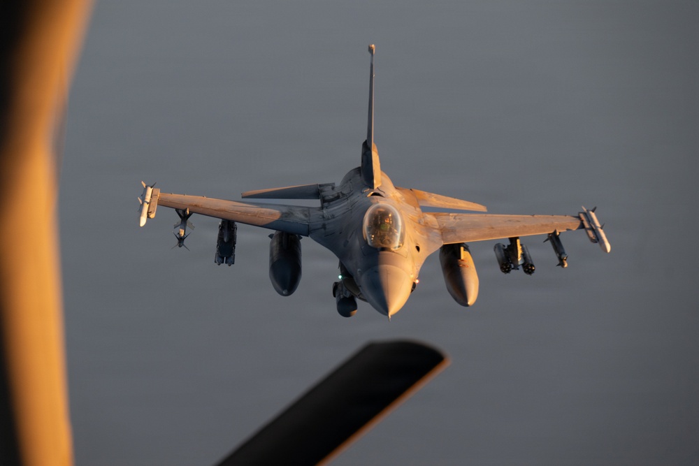 A front-facing view of an F-16 Fighting Falcon jet fighter aircraft in flight, photographed from what appears to be a tanker aircraft during an aerial refueling approach. The aircraft is captured in warm golden light consistent with sunrise or sunset conditions. The jet is carrying multiple munitions and external stores on its wing hardpoints.