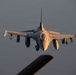 A front-facing view of an F-16 Fighting Falcon jet fighter aircraft in flight, photographed from what appears to be a tanker aircraft during an aerial refueling approach. The aircraft is captured in warm golden light consistent with sunrise or sunset conditions. The jet is carrying multiple munitions and external stores on its wing hardpoints.