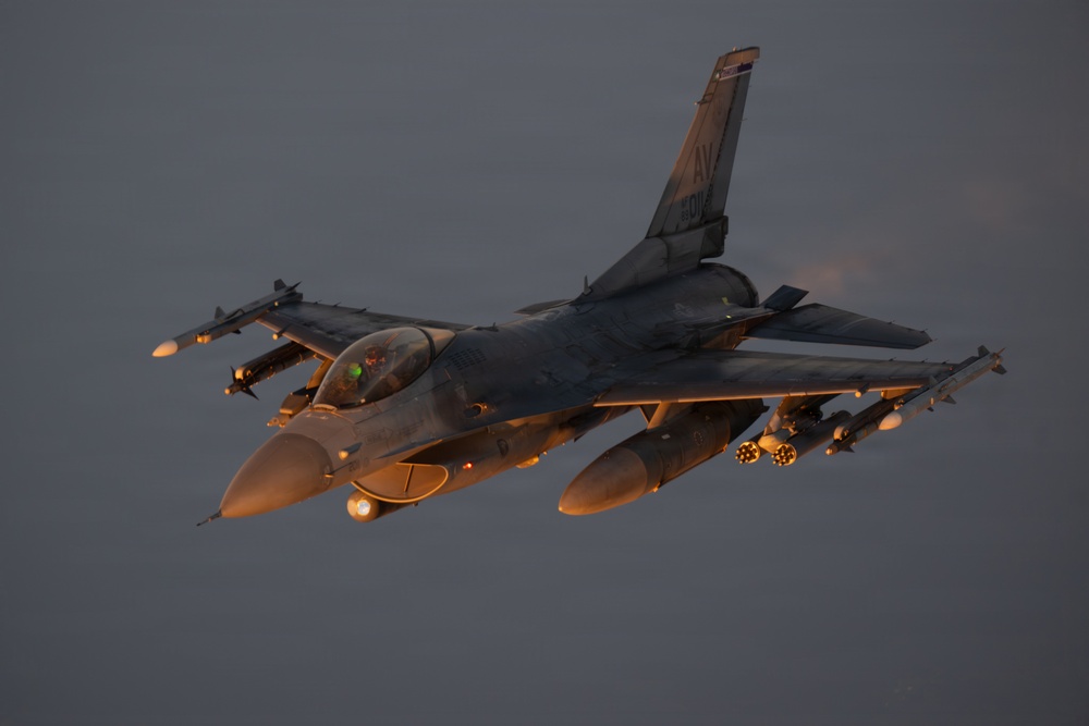 A U.S. Air Force F-16 Fighting Falcon is captured in flight during low-light conditions, likely at dusk or dawn. The aircraft is heavily loaded with multiple munitions and external stores on its wing pylons. The tail code 'AV' is visible, indicating assignment to Aviano Air Base, Italy.