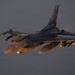A U.S. Air Force F-16 Fighting Falcon is captured in flight during low-light conditions, likely at dusk or dawn. The aircraft is heavily loaded with multiple munitions and external stores on its wing pylons. The tail code 'AV' is visible, indicating assignment to Aviano Air Base, Italy.