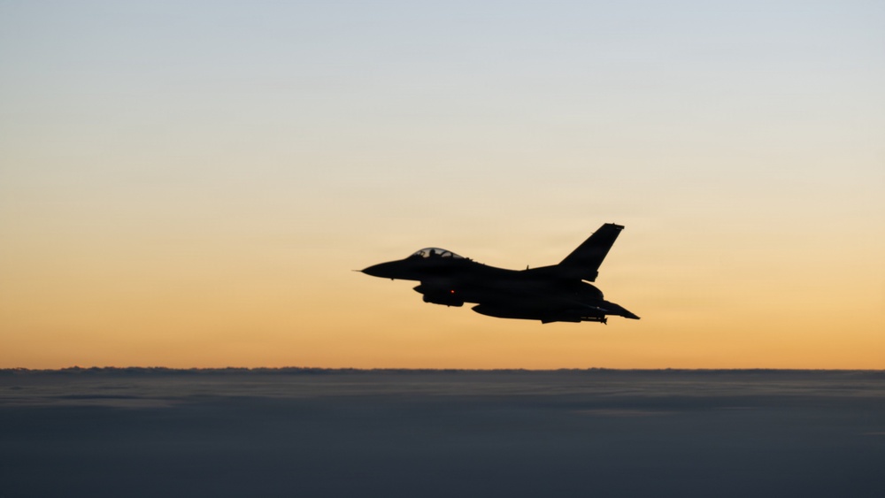 A military fighter jet, consistent with an F-16 Fighting Falcon silhouette, is captured in flight above a cloud layer during twilight or dusk conditions. The aircraft is rendered almost entirely in silhouette against a warm orange-to-blue gradient sky. A single red navigation light is visible on the underside of the aircraft.