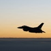 A military fighter jet, consistent with an F-16 Fighting Falcon silhouette, is captured in flight above a cloud layer during twilight or dusk conditions. The aircraft is rendered almost entirely in silhouette against a warm orange-to-blue gradient sky. A single red navigation light is visible on the underside of the aircraft.