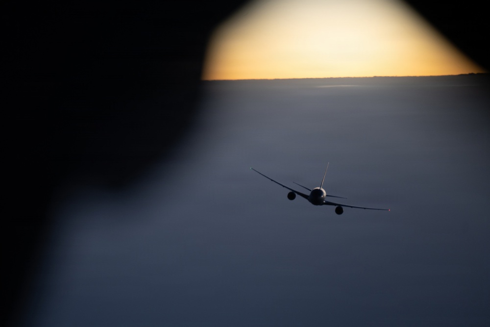 A commercial jet aircraft is photographed in flight at high altitude during sunset or sunrise, as seen through what appears to be another aircraft's window. The aircraft is silhouetted against a dramatic sky with a golden horizon and a vast cloud layer below. The photo was taken from a nearby aircraft, suggesting formation flying or close proximity flight operations.