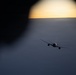 A commercial jet aircraft is photographed in flight at high altitude during sunset or sunrise, as seen through what appears to be another aircraft's window. The aircraft is silhouetted against a dramatic sky with a golden horizon and a vast cloud layer below. The photo was taken from a nearby aircraft, suggesting formation flying or close proximity flight operations.