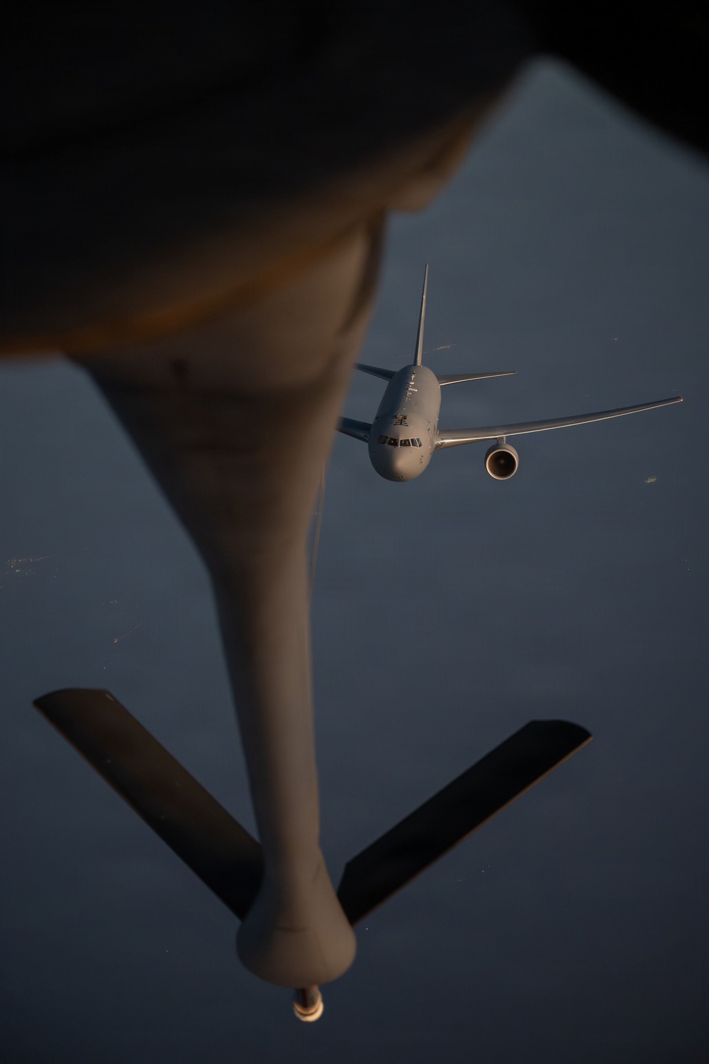 An aerial refueling operation is captured from the perspective of a tanker aircraft looking down its refueling boom toward a receiver aircraft approaching from below. The receiver aircraft appears to be a large military or military-derivative wide-body jet, visible head-on against a hazy blue sky background. The refueling boom with its ruddervator control surfaces is prominently visible in the foreground.