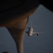 An aerial refueling operation is captured from the perspective of a tanker aircraft looking down its refueling boom toward a receiver aircraft approaching from below. The receiver aircraft appears to be a large military or military-derivative wide-body jet, visible head-on against a hazy blue sky background. The refueling boom with its ruddervator control surfaces is prominently visible in the foreground.