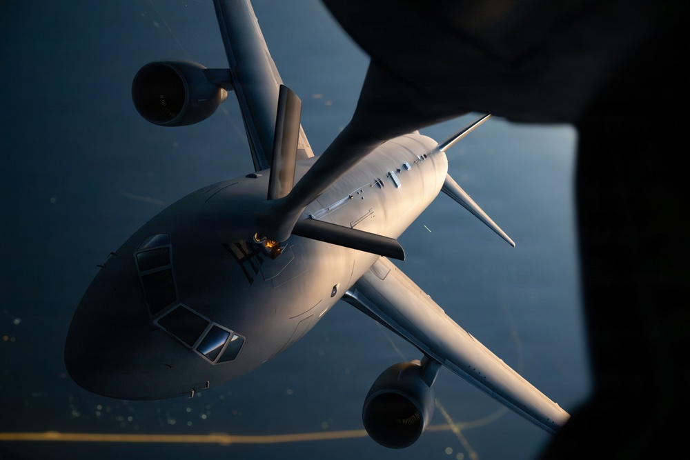 An aerial photograph taken from above showing a large military tanker aircraft in flight, viewed from the perspective of another aircraft above it. The image captures what appears to be an aerial refueling operation, with the tanker aircraft positioned below and slightly forward of the camera aircraft. Warm golden light illuminates the top surfaces of the aircraft against a dark blue background suggesting high altitude flight over water or terrain.