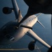 An aerial photograph taken from above showing a large military tanker aircraft in flight, viewed from the perspective of another aircraft above it. The image captures what appears to be an aerial refueling operation, with the tanker aircraft positioned below and slightly forward of the camera aircraft. Warm golden light illuminates the top surfaces of the aircraft against a dark blue background suggesting high altitude flight over water or terrain.